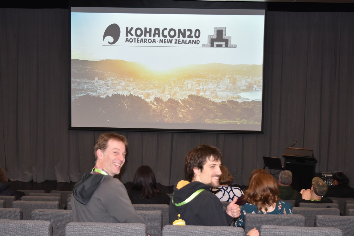 Two attendees at Kohacon turning to face the camera and smiling with the projector screen behind them showing Kohacon's logo