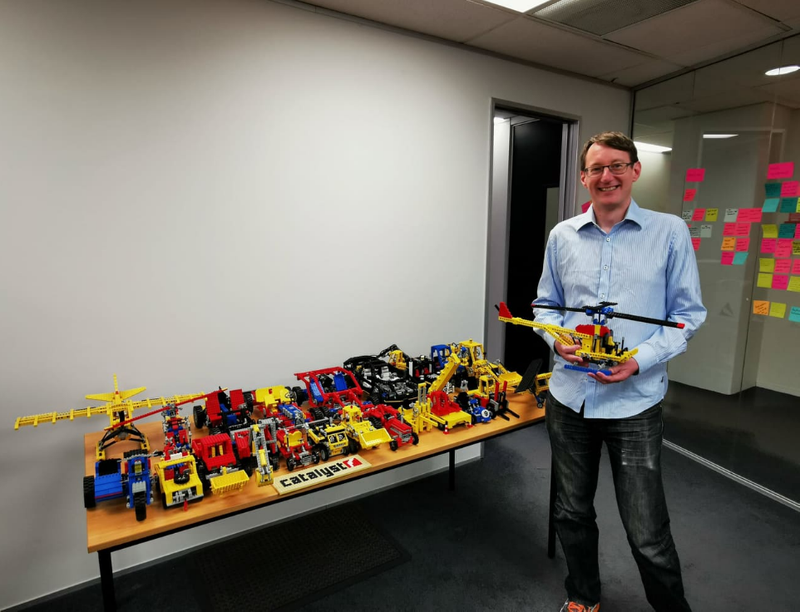 A person is standing in an office holding a Lego helicopter, next to a table with many other Lego sets on it.