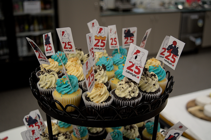 A lot of cupcakes with blue or white icing and sprinkles and flags saying Catalyst 25.