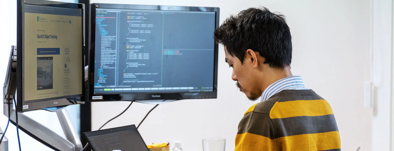 A person is sitting at a desk looking at two computer monitors with code on the screens.