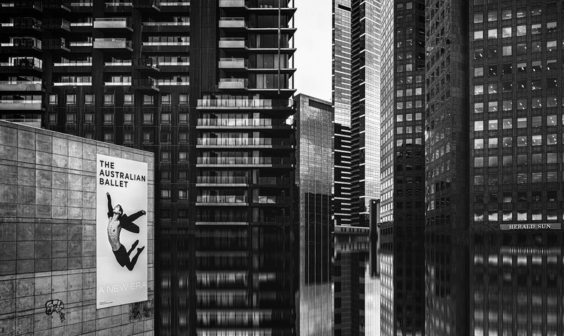 A black and white photo of tall city buildings with reflections in the shiny street below, and in the foreground is a large poster of a ballet dancer in midair.