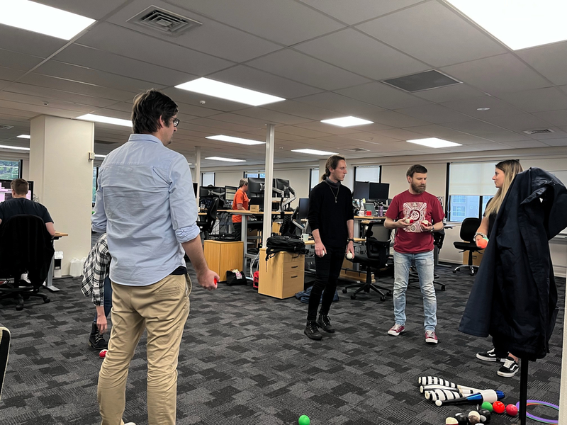 A handful of people are standing in an office talking, and holding various things to juggle.