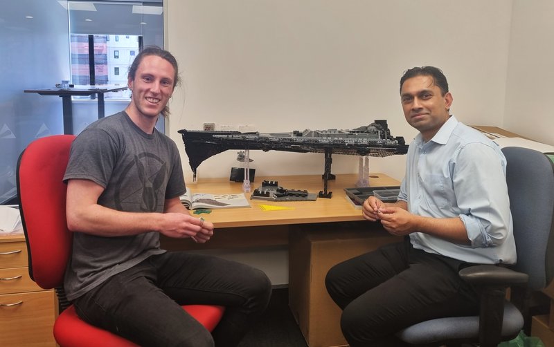 Liam Sharpe and Sanjay D&#x27;Souza are sitting on chairs in a brightly lit office with a large, unfinished Star Wars Lego set on the table behind them.