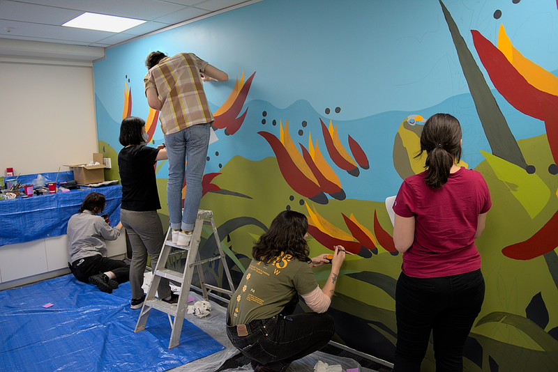Several people, including one person on a ladder, are painting a mural on a wall in a meeting room with bright blue, green, red, and yellow colours.
