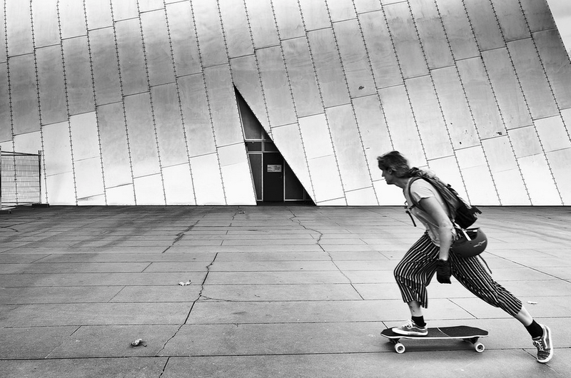 A black and white image of a skater with a facade in the background that has a triangle shape, reflected in the skater&#x27;s posture.