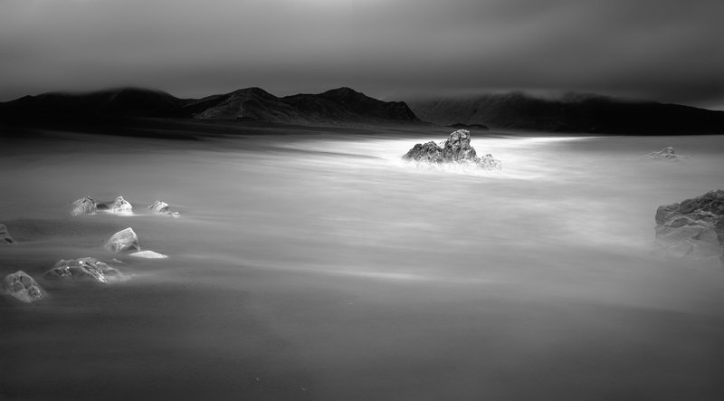 A black and white landscape photo with a line of black hills in the background and blurry water around rocks in the foreground.